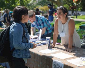 A student talks to a woman over a table outside with information pamphlets strewn across it.