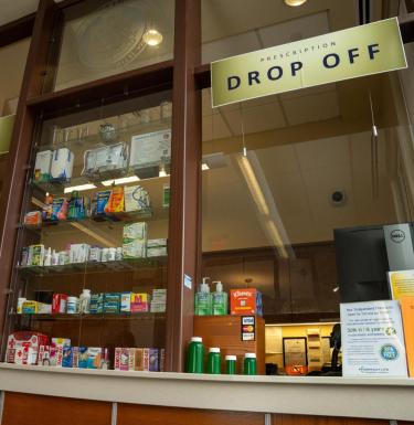 Pharmacy window with shelves of various pill bottles and a window with a sign that reads "drop off."