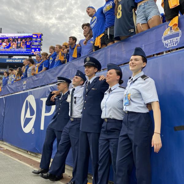 Group of five cadets in uniform standing in front of a group of students.