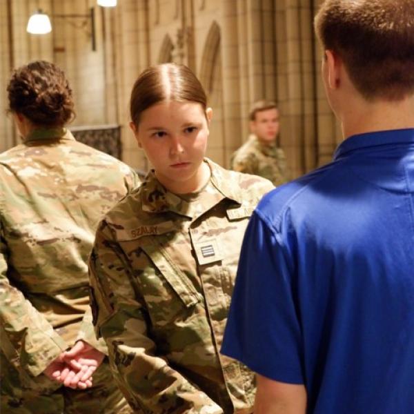 A woman in a military uniform inspecting the outfit of a man in a blue polo shirt.