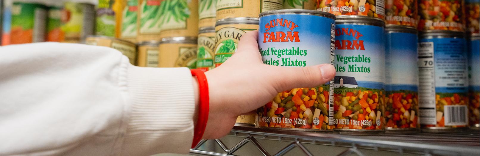A hand reaching for a can of mixed vegetables.