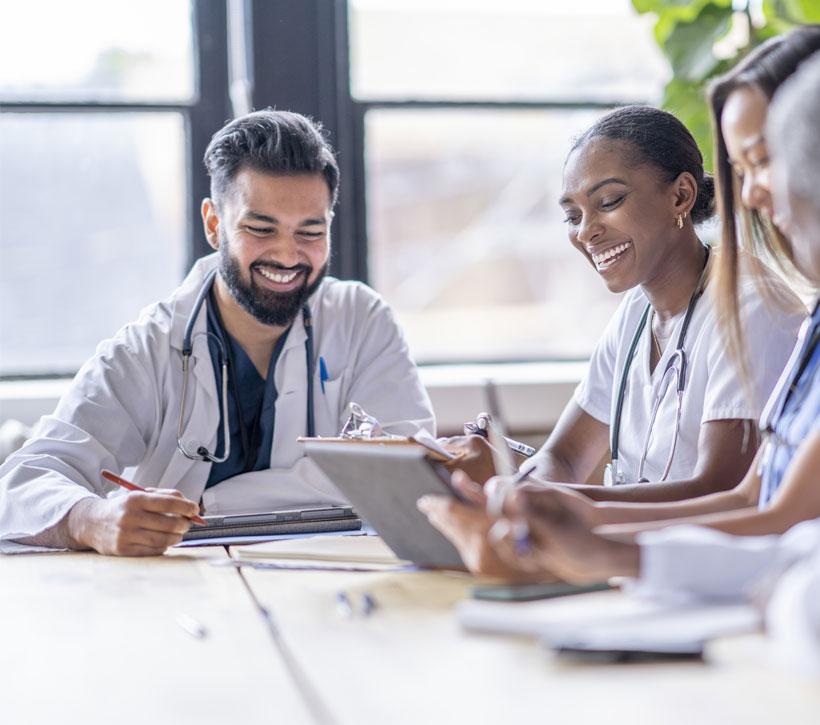 A smiling man and woman in white coats look at papers and a laptop on a table.