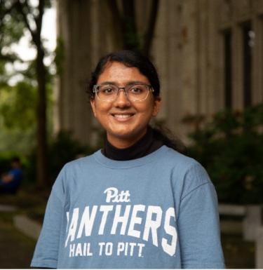 Woman with long dark hair in a ponytail with glasses smiling in a light blue crewneck sweatshirt.