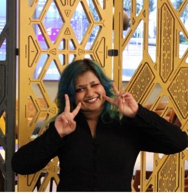 Person with medium length brown and teal hair smiling and holding up two peace signs with their hands.