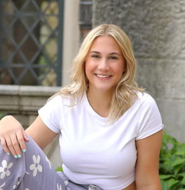 Blonde woman in a white t-shirt and purple jeans sits on the ground and smiles.