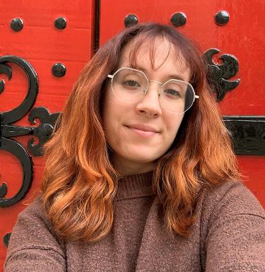 A woman with light brown hair standing in front of a red door in a brown sweater smiling.