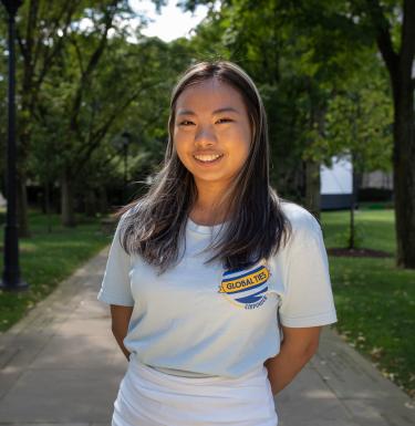 Outdoor headshot of a smiling woman with long dark hair in a Global Ties tee shirt.