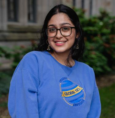 Outdoor headshot of a smiling woman in dark square glasses with dark hair in a blue Global Ties crewneck shirt.