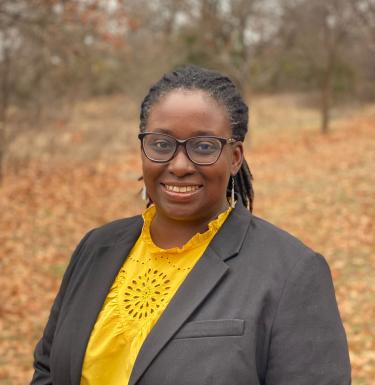 A woman standing outside smiling in a yellow blouse and grey blazer.