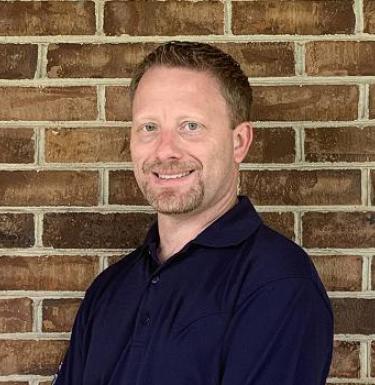 Man in a dark sweater smiling in front of a brick wall.