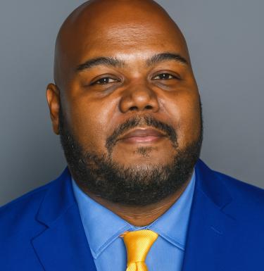 A photo of a bald man with facial hair in a blue suit.