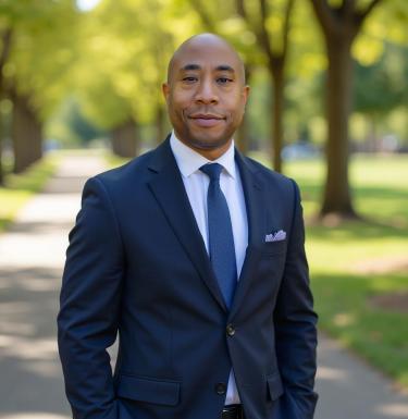 A man standing and smiling in a navy suit in front of trees.