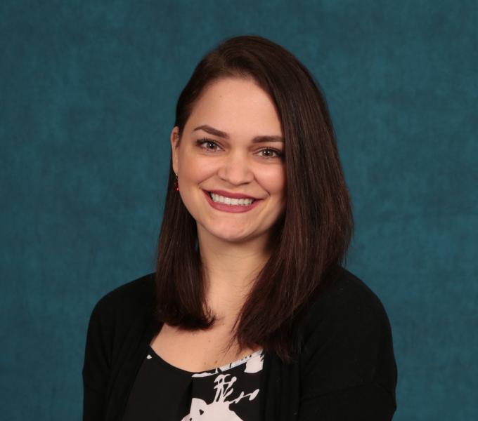 A woman with short dark hair in a black sweater and floral top smiling in front of a green backdrop.