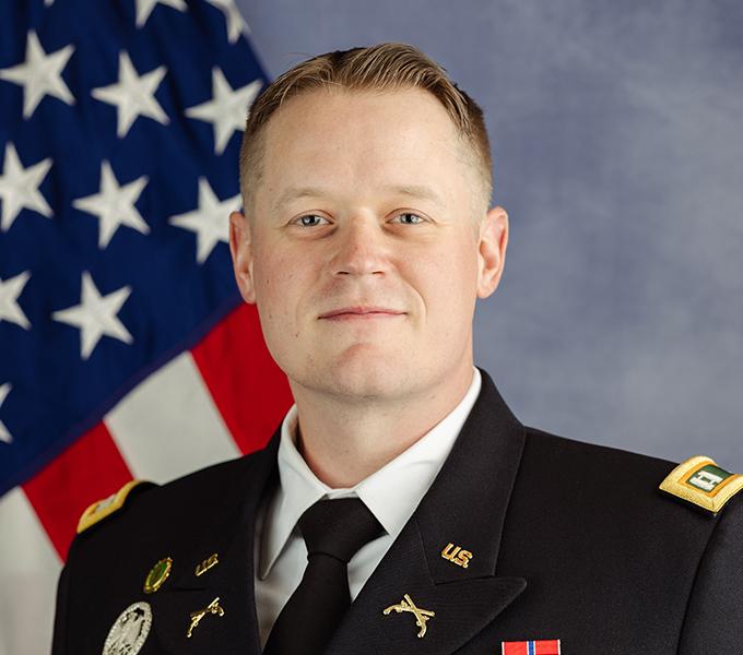 Photo of a man in a military uniform with blonde hair smiling in front of a grey background.