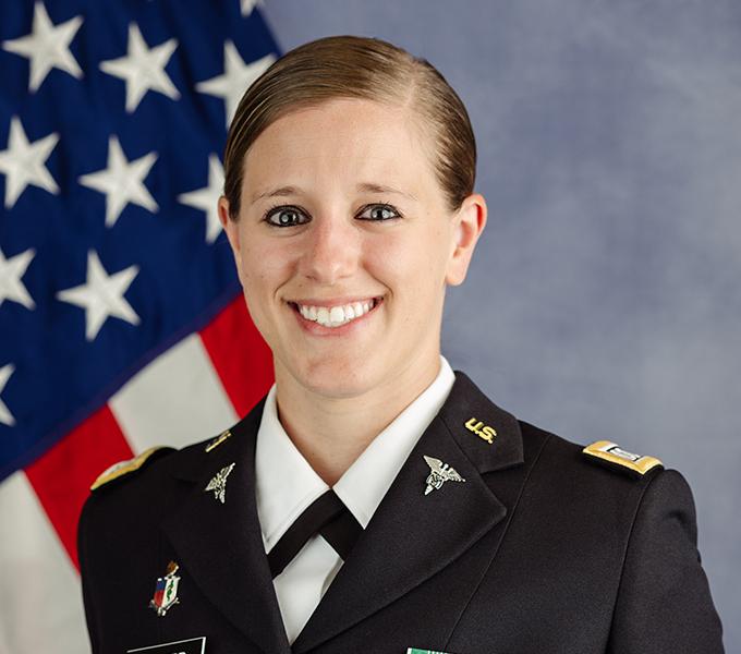 A woman in a military uniform with blonde hair smiling in front of a grey background.