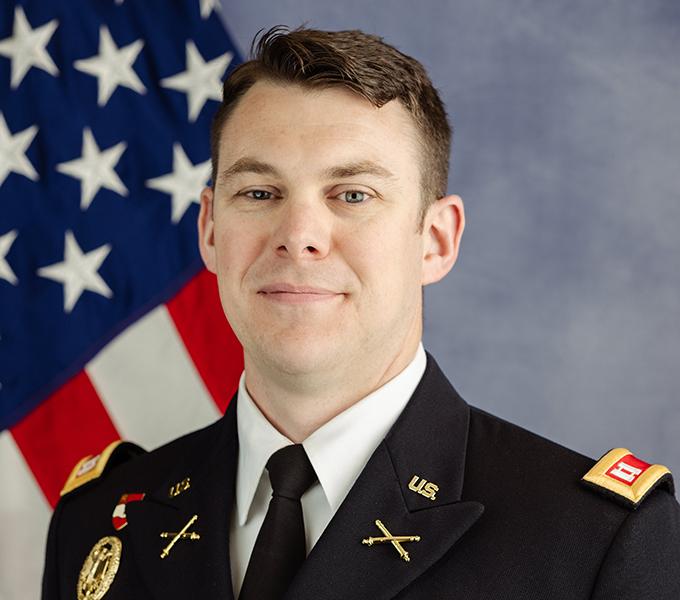 Photo of a man in a military uniform with dark hair smiling in front of a grey background.