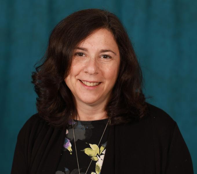 A woman with dark curly hair smiling in front of a green backdrop in a black sweater and floral top.