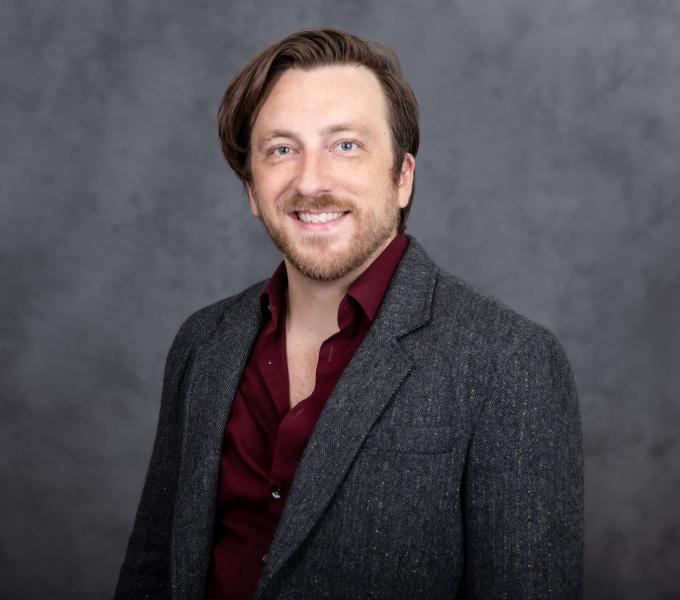 Headshot of a man with swooping brown hair in a gray cardigan and maroon shirt.