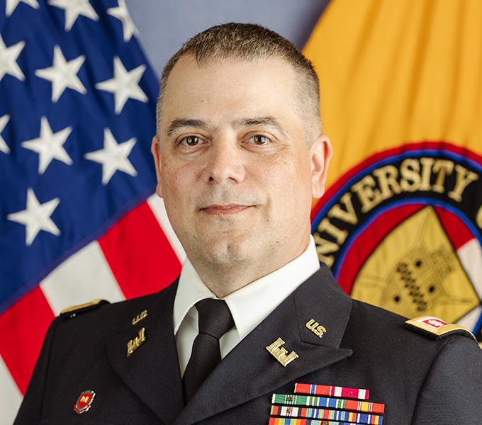 Photo of a man in a military uniform with dark hair smiling in front of a grey background.