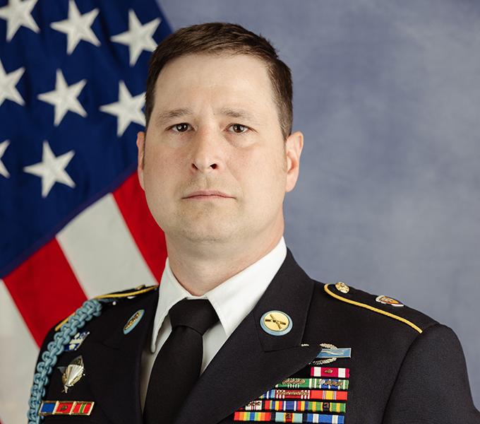 Photo of a man in a military uniform with dark hair smiling in front of a grey background.