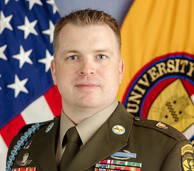 Photo of a man in a military uniform with blonde hair smiling in front of a grey background.