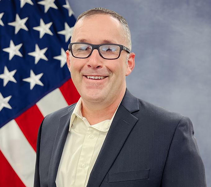 A man smiling in a grey suit with glasses smiling in front of a grey background.