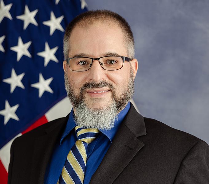 Photo of a man in a black suit with dark hair and a grey beard smiling in front of a grey background.